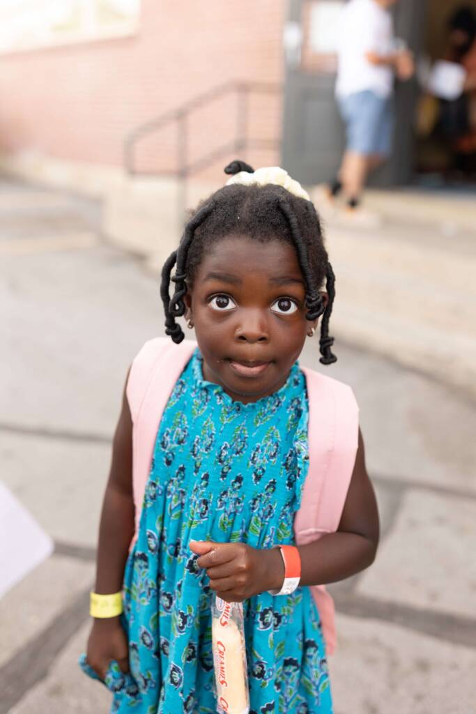 A young girl wearing a pink backpack looks up curiously while holding a snack at Utah Refugee Connection’s Back to School event.