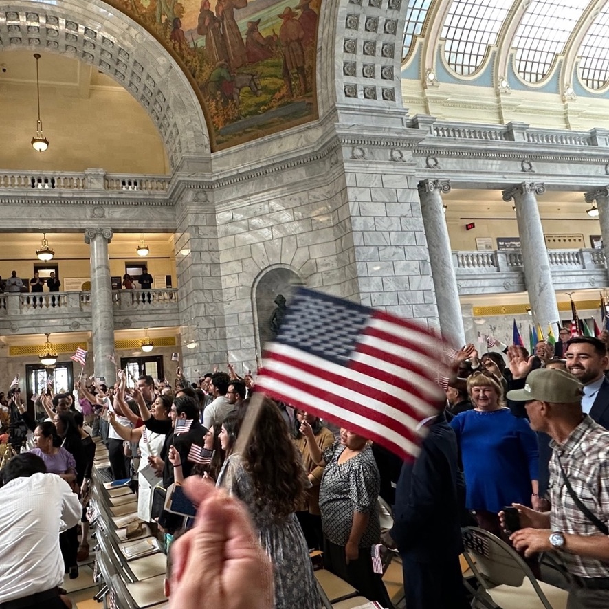 Community members wave American flags inside a historic building during a naturalization ceremony, celebrating refugees in Utah as they become citizens and fully join their new communities.