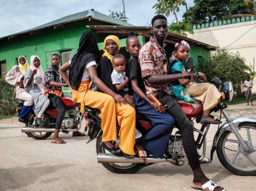 A group of displaced people, including children and adults, share a crowded motorcycle in a community setting, reflecting the resilience of refugee families and the global context behind the refugee stories Utah Refugee Connection seeks to honor and support.
