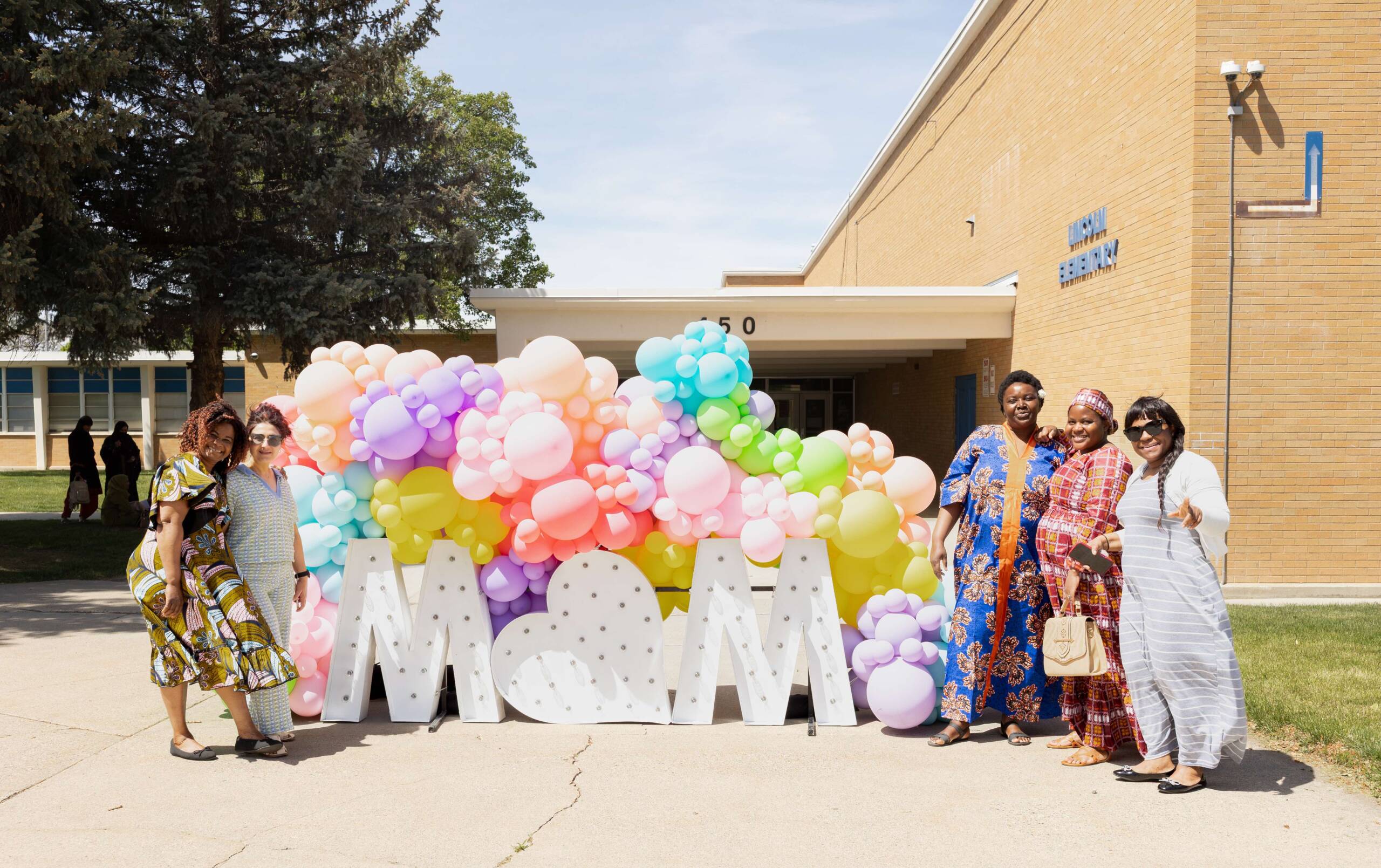 Refugee mothers pose beside a colorful MOM display during Utah Refugee Connection’s Mother’s Day celebration in Utah. The event celebrated refugee mothers with joy, community connection, and support.